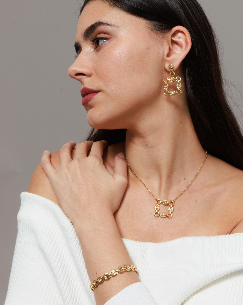 Woman wearing gold jewelry including earrings, necklace, and bracelet against a neutral background