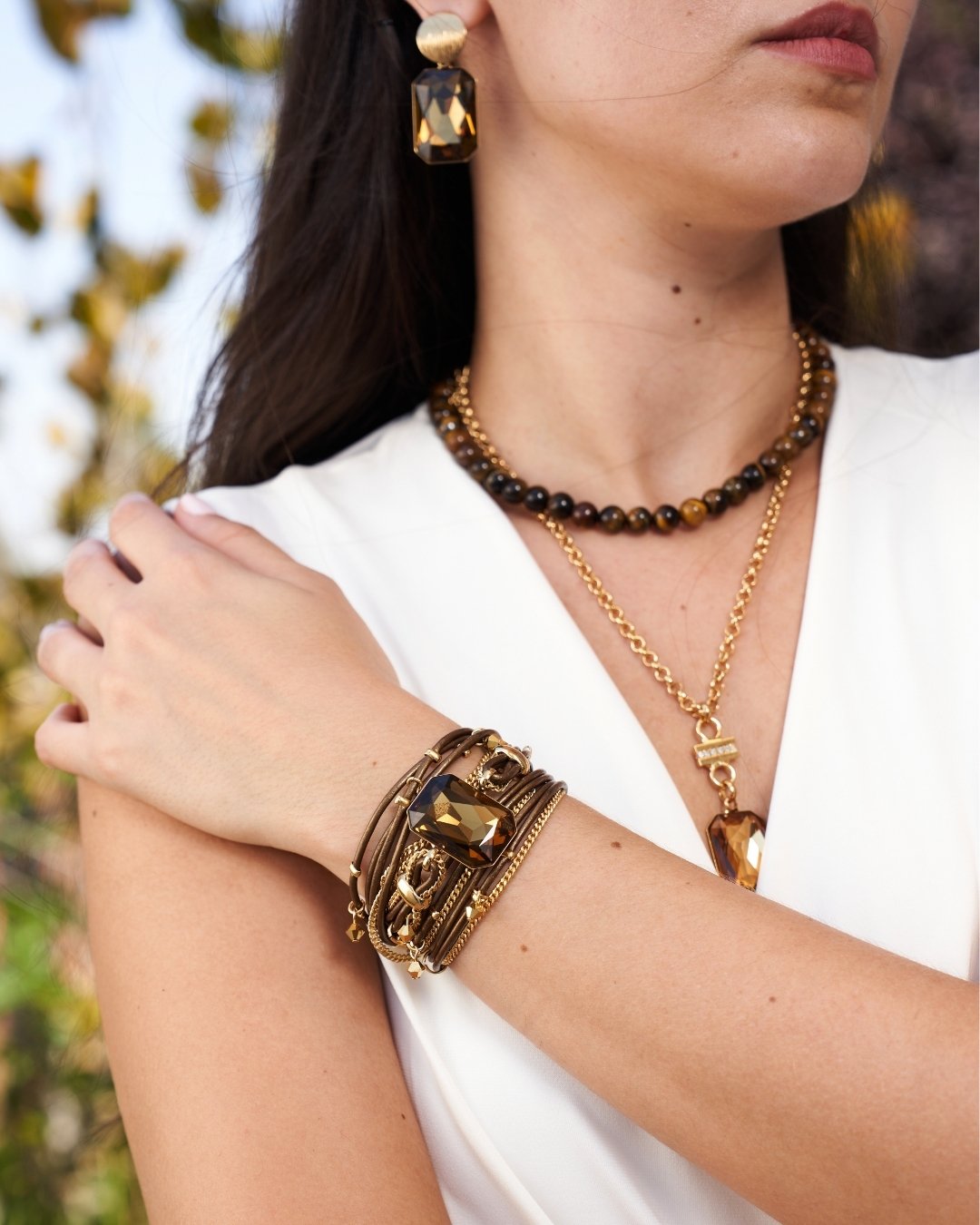 A close-up lifestyle shot of a woman wearing a gold-toned multi-band cuff bracelet with a faceted brown gemstone, along with layered necklaces and earrings.