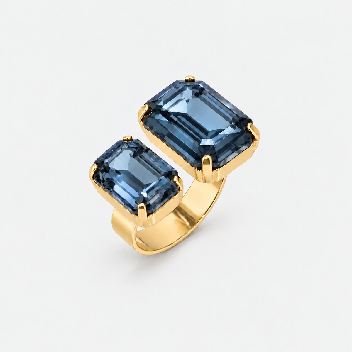 A gold ring featuring two rectangular emerald-cut blue gemstones in separate prong settings, shown on a light gray background.
