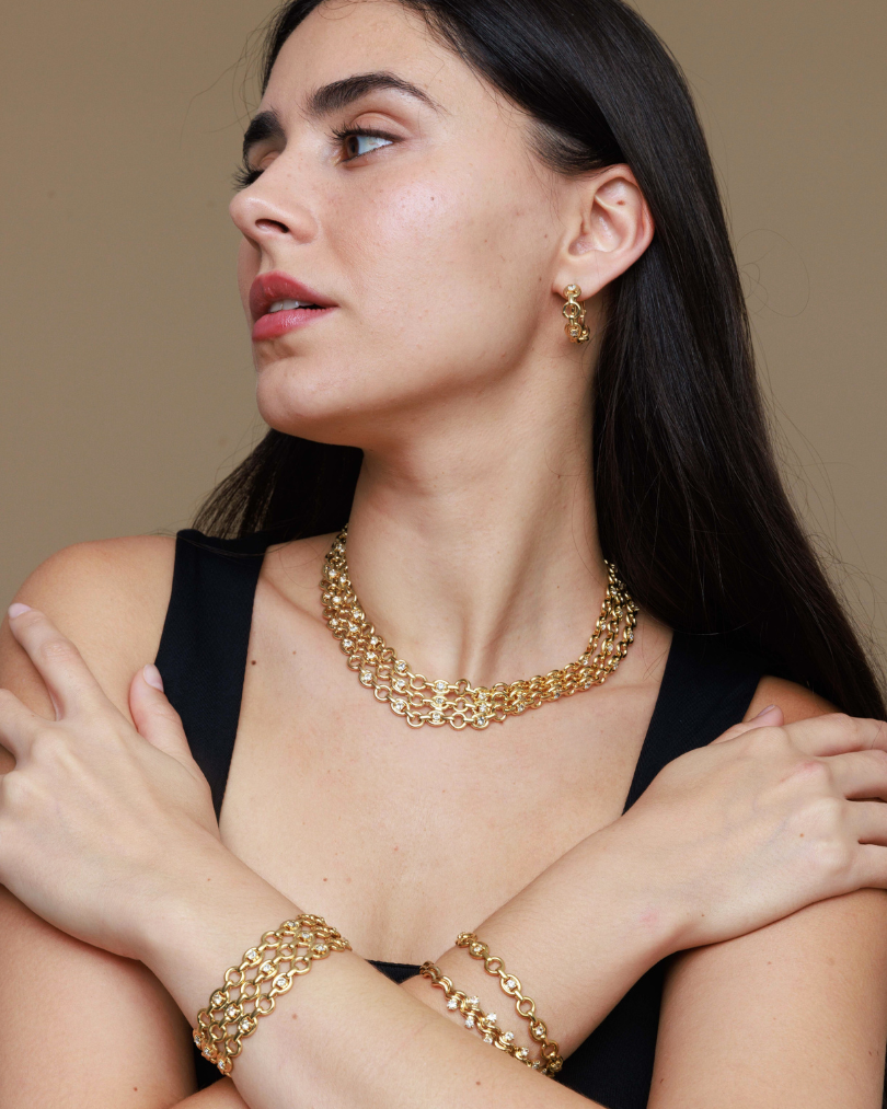 Woman wearing gold jewelry including a necklace, bracelet, and earrings against a neutral background