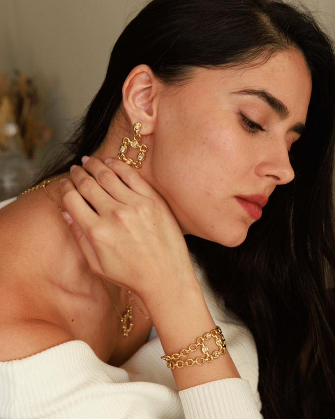 Woman wearing gold chain earrings and bracelet, close-up of face and hand.