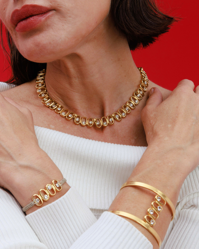 Woman wearing gold jewelry including a necklace, bracelet, and ring against a red background