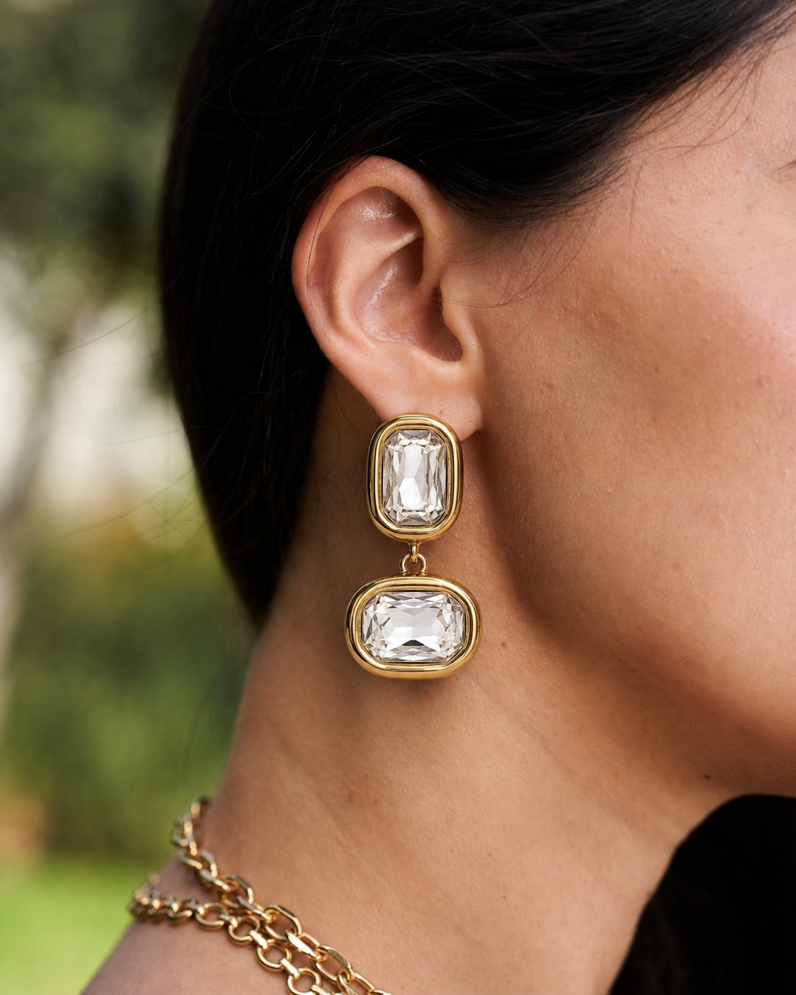A close-up of a model's ear wearing a gold drop earring with two rectangular clear gemstones, set against a blurred outdoor background.