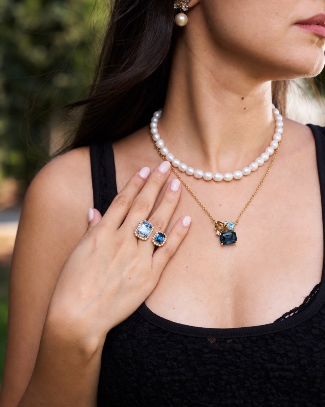 A close-up lifestyle shot of a woman wearing a layered jewelry set, featuring a white pearl necklace, a gold chain with a dark blue pendant, and matching blue and gold stud earrings, while wearing a black lace top.