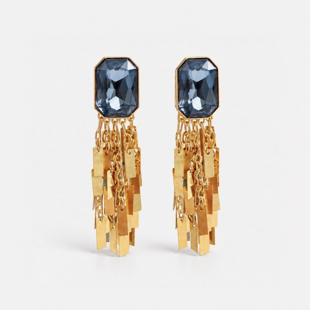 A pair of gold dangle earrings featuring faceted dark blue-gray rectangular gemstones at the top and multiple dangling gold-tone chain and rectangular charms below, shown on a light gray background.