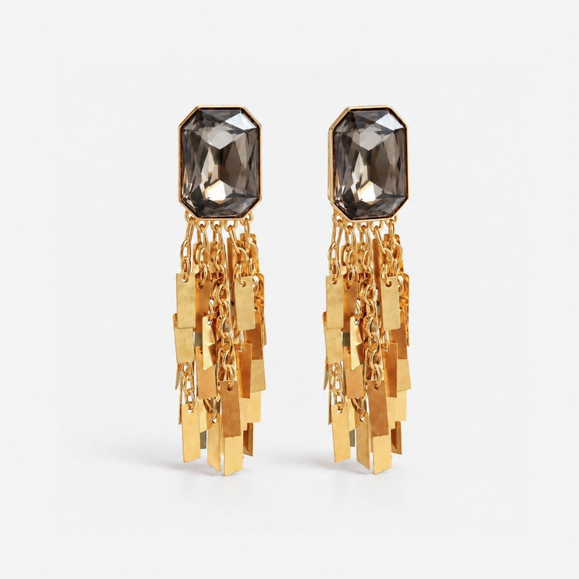 A pair of gold dangle earrings with a faceted smoky gray stone at the top and multiple dangling gold-tone chain and rectangular charms, shown on a light gray background.