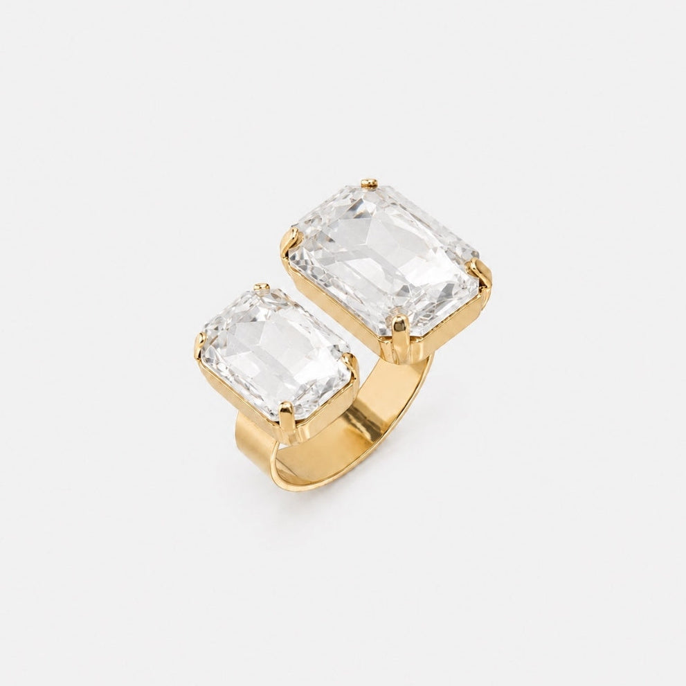 A gold ring featuring two large, clear rectangular emerald-cut gemstones in a split setting, photographed on a clean, light gray background.