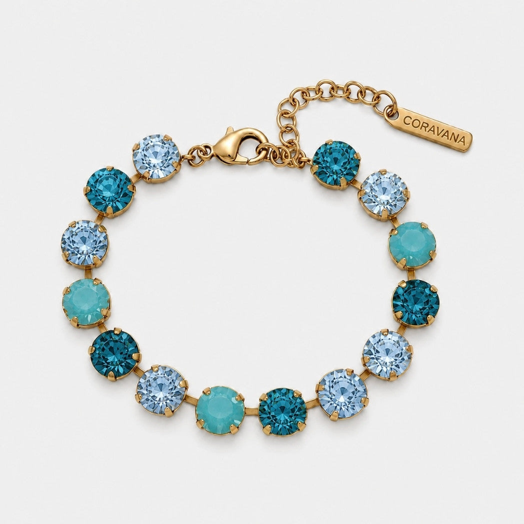A gold-tone bracelet with alternating round light blue and teal-blue rhinestones, featuring a lobster clasp, extension chain, and a branded tag that reads CORAVANA.