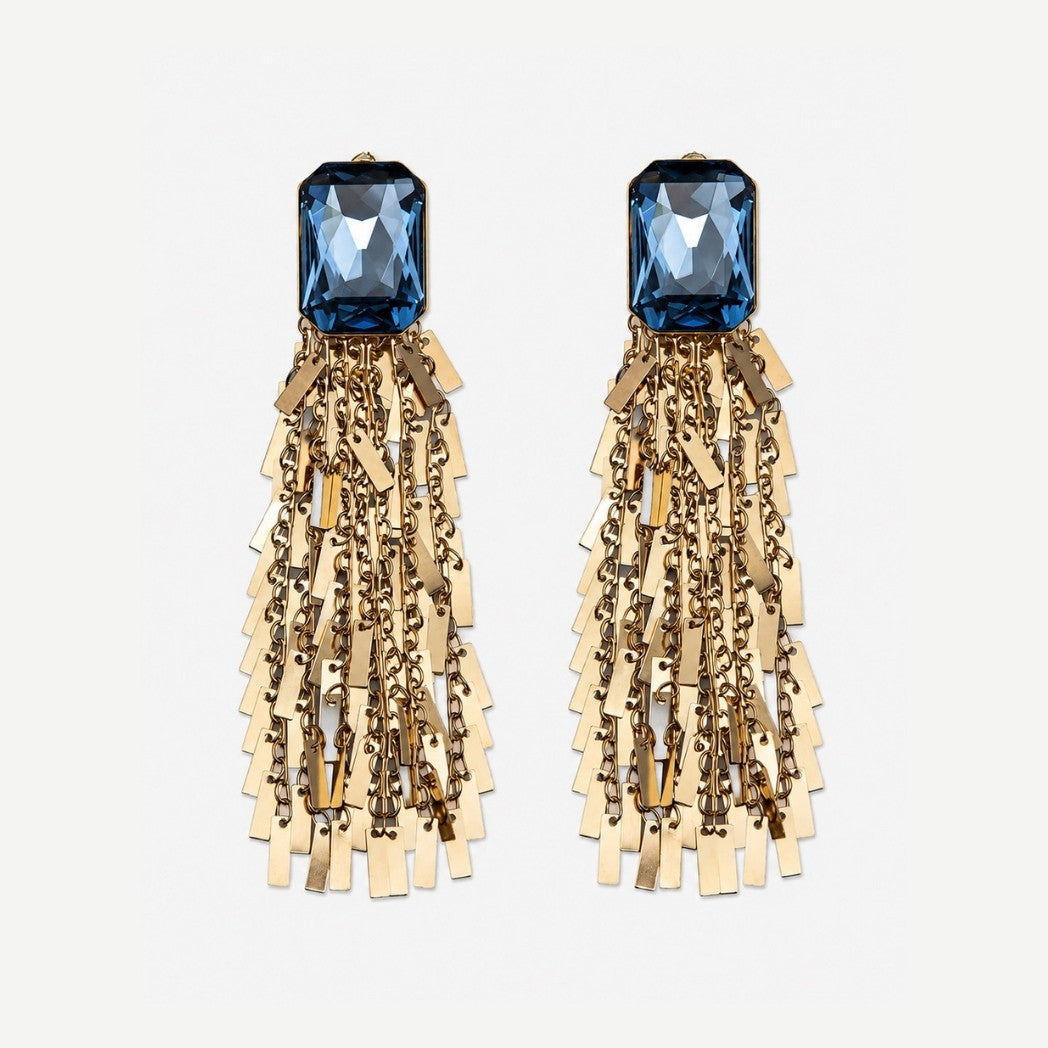 A pair of statement dangle earrings featuring a faceted blue gemstone at the top, with cascading gold-toned chains and rectangular metal fringe below, set against a plain light gray background.