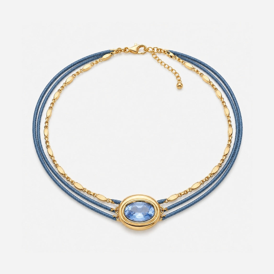 A multi-strand necklace featuring blue cord and gold chain, with a central oval gold bezel holding a light blue gemstone, shown on a light gray background.