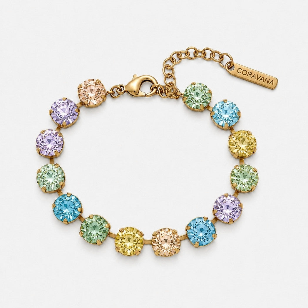 A gold-plated bracelet with multiple round, bezel-set crystals in pastel shades of blue, green, yellow, and purple, shown on a clean white background. A small tag on the clasp chain reads CORAVANA.