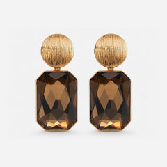 The Sunburst Earrings, gold-tone drop earrings with a round textured sunburst stud and a faceted rectangular gemstone drop, on a light gray background.