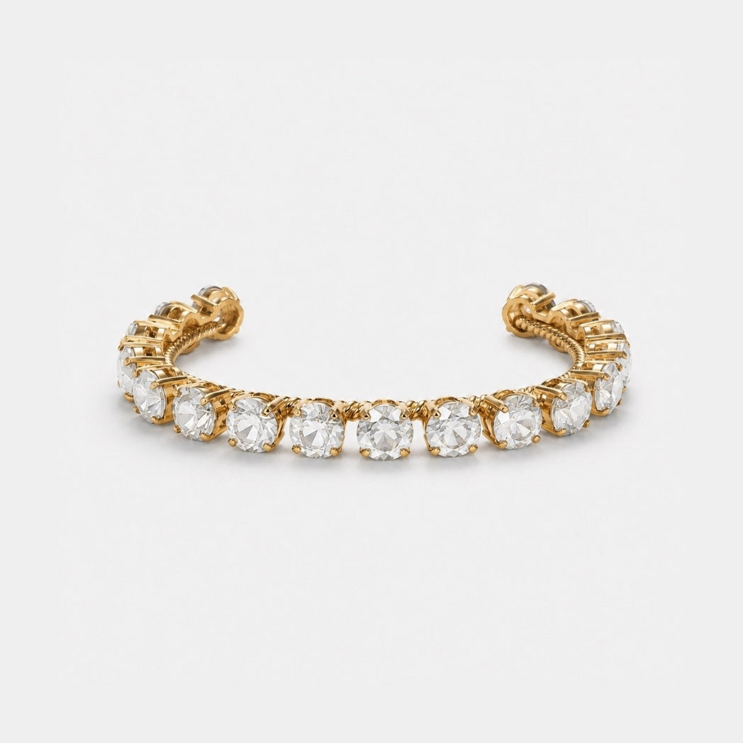A gold-toned cuff bracelet featuring a row of round clear gemstones in a prong setting, photographed against a clean light gray background.