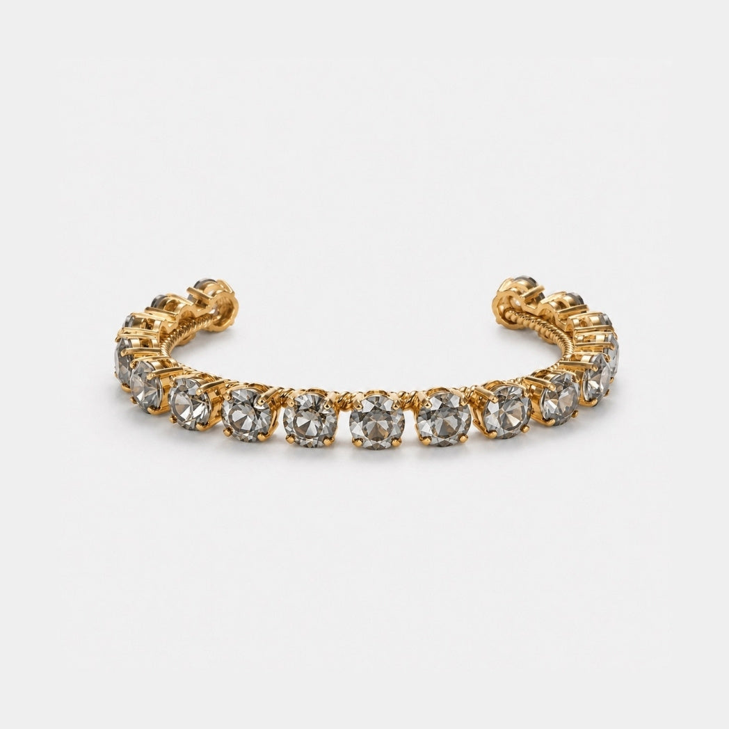 A gold-toned cuff bracelet featuring a row of round clear gemstones, photographed on a clean light gray background.