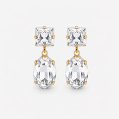 A pair of gold drop earrings featuring a square clear crystal stud and a larger oval clear crystal dangle, set in gold on a light gray background.