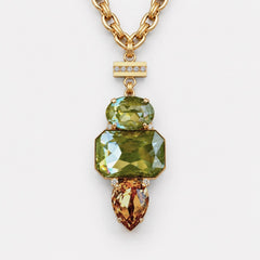 A gold-tone pendant necklace featuring a large faceted green gemstone, a smaller oval green gemstone, and a pear-cut amber gemstone, suspended from a gold chain on a light gray background.
