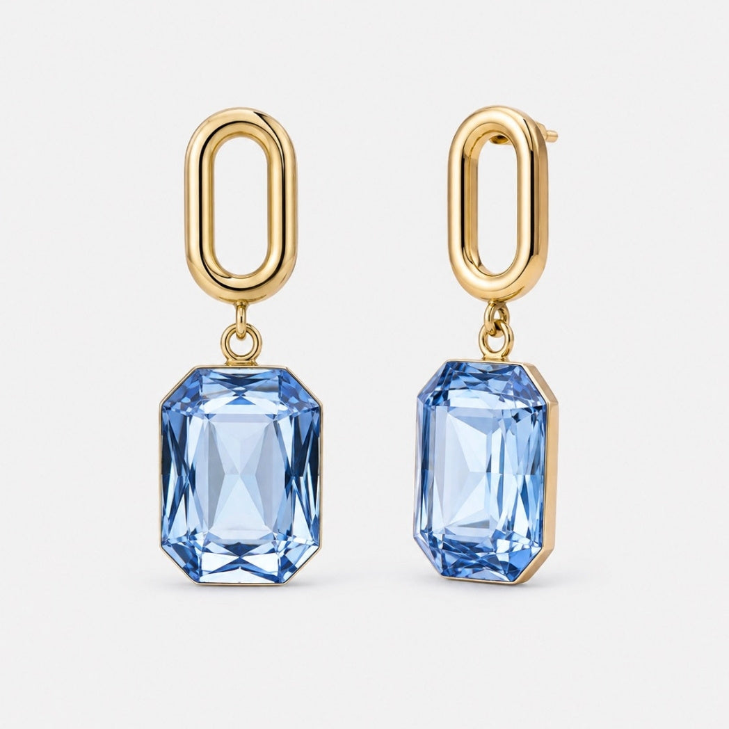 A pair of gold drop earrings featuring faceted light blue rectangular gemstones, shown against a clean, light gray background.