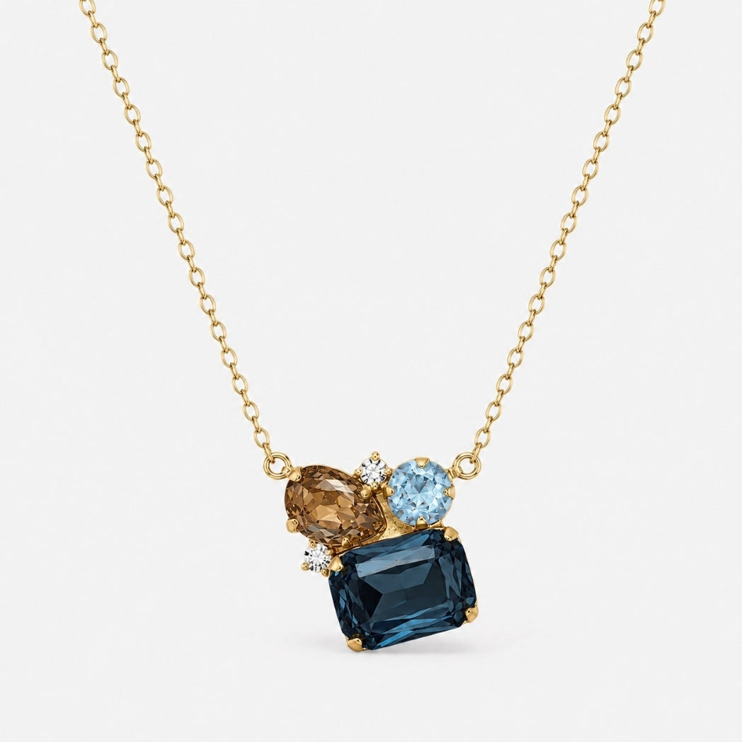 A gold chain necklace featuring a multi-stone pendant with a large rectangular dark blue gemstone, an oval champagne-colored gemstone, and a round light blue gemstone, set against a light gray background.