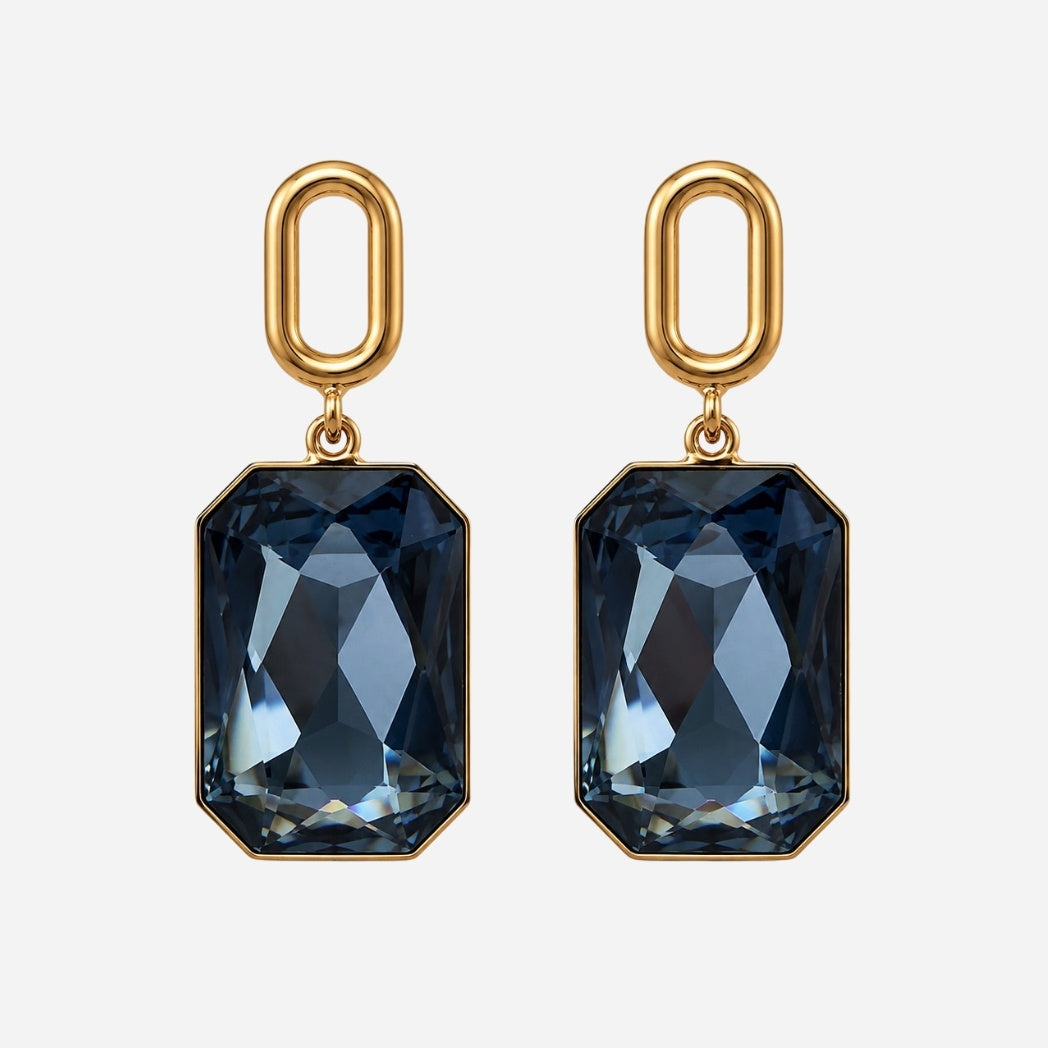 A pair of gold drop earrings featuring faceted dark blue-gray rectangular gemstones, shown against a light gray background.