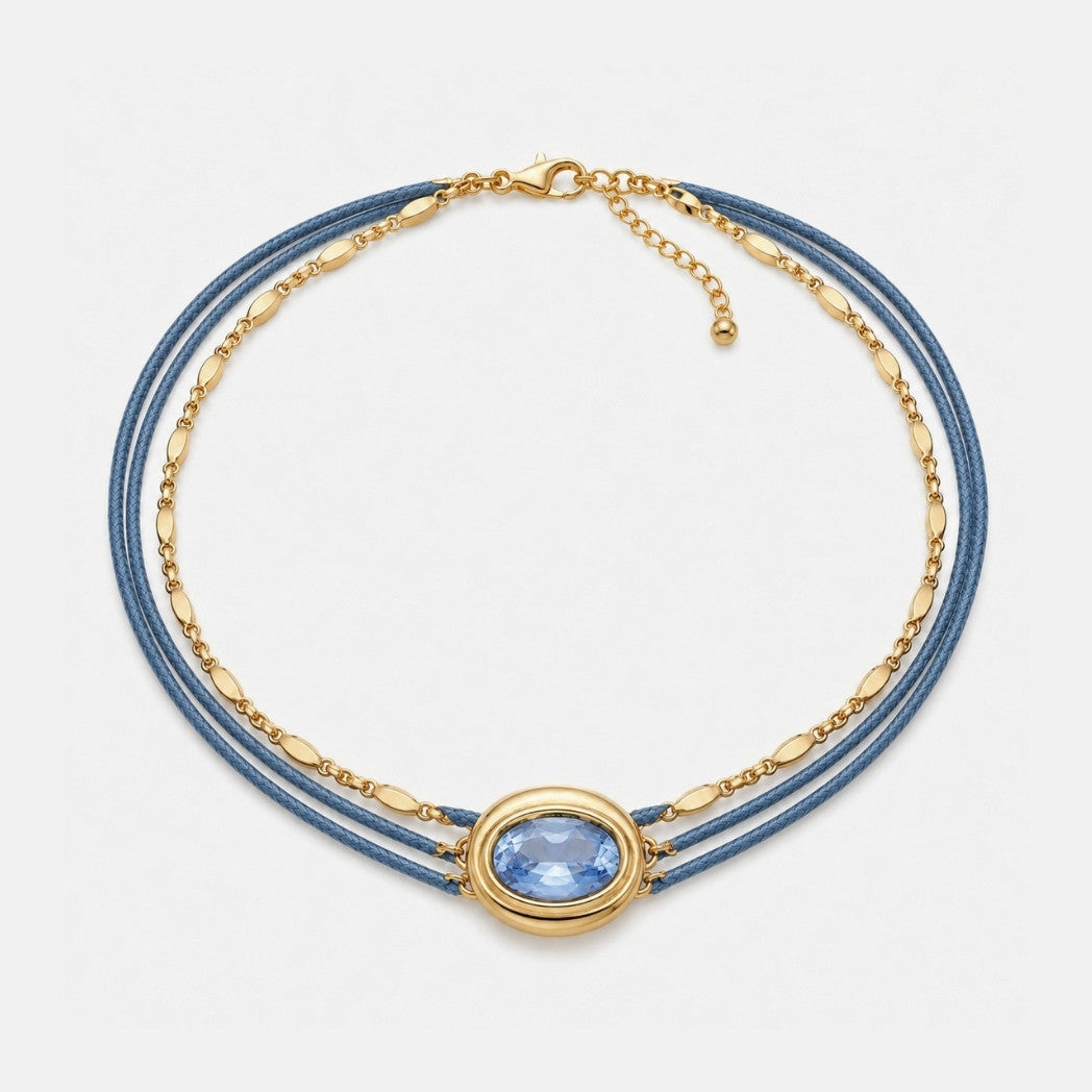 A multi-strand necklace featuring blue cords, a gold chain, and a central oval gold bezel holding a light blue gemstone, displayed on a clean white background.