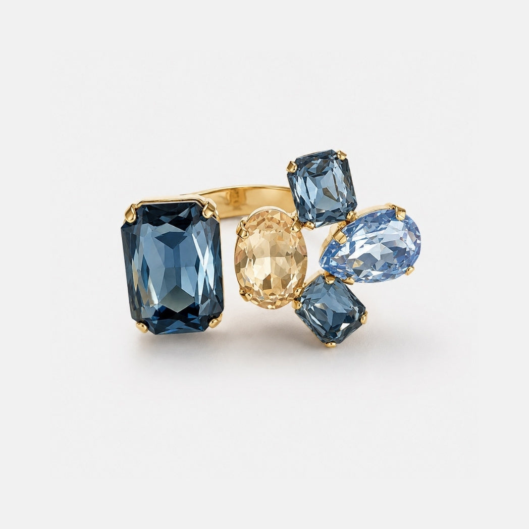 A gold ring featuring multiple blue gemstones of different shapes (rectangular, oval, pear, and round) and one small champagne-colored oval stone, set against a clean white background.