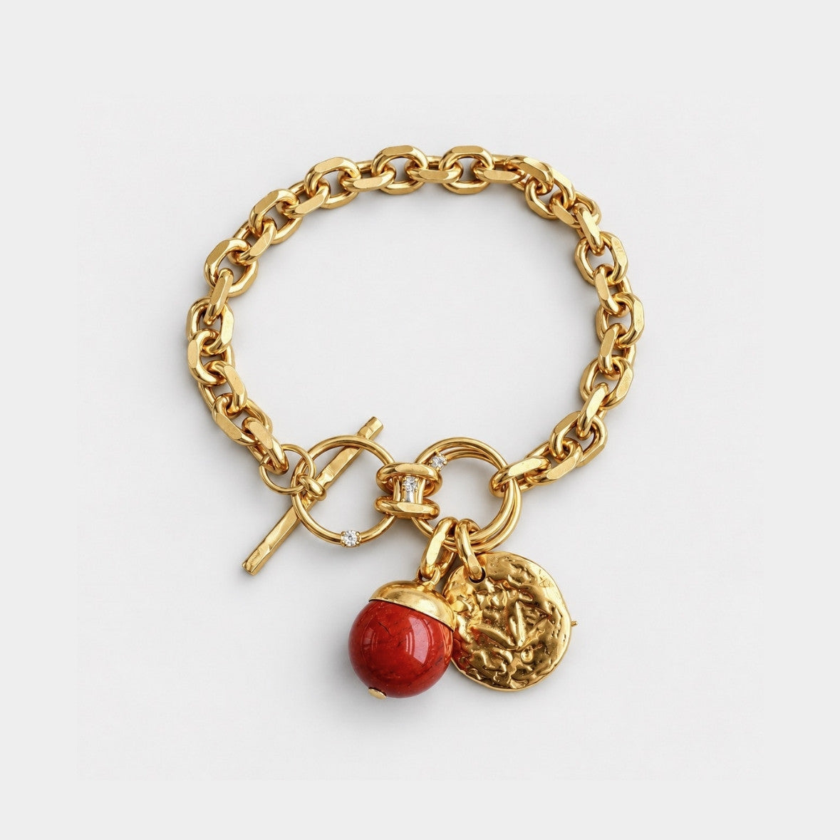 A gold chain bracelet with a toggle clasp, featuring two dangling charms: a polished red bead and a textured gold coin charm.