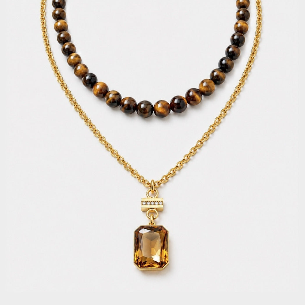A double-strand gold necklace set on a light gray background. The top strand features round brown and black tiger's eye beads, while the bottom strand is a gold chain with a large faceted amber gemstone pendant and a small gold bar with tiny clear stones.