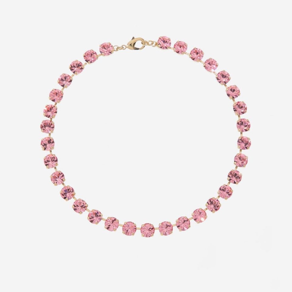 A delicate tennis-style necklace featuring small, round-cut pink crystals set in a gold-tone metal, displayed against a clean, light gray background.