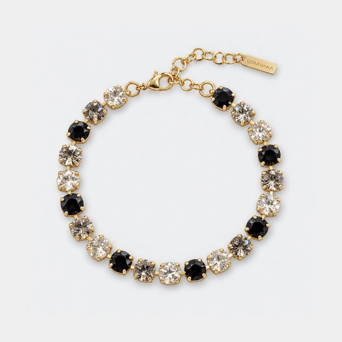 A gold-tone tennis bracelet featuring alternating round black and clear crystals, displayed on a clean white background. A small gold tag on the clasp chain reads 'ORAGNA'.