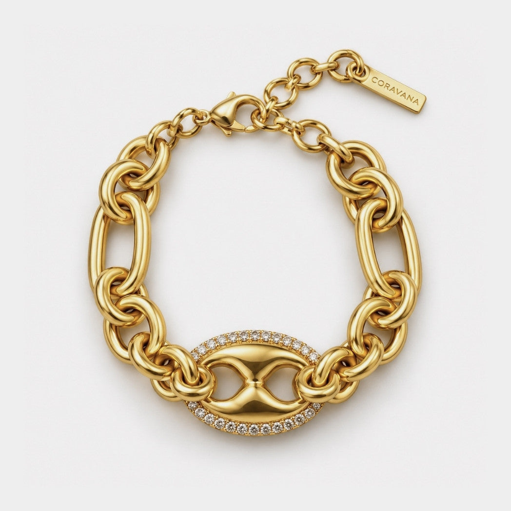 A gold-tone chain bracelet with a central hourglass motif paved with small clear stones, shown on a clean white background. A small tag on the chain reads CORAVANA.