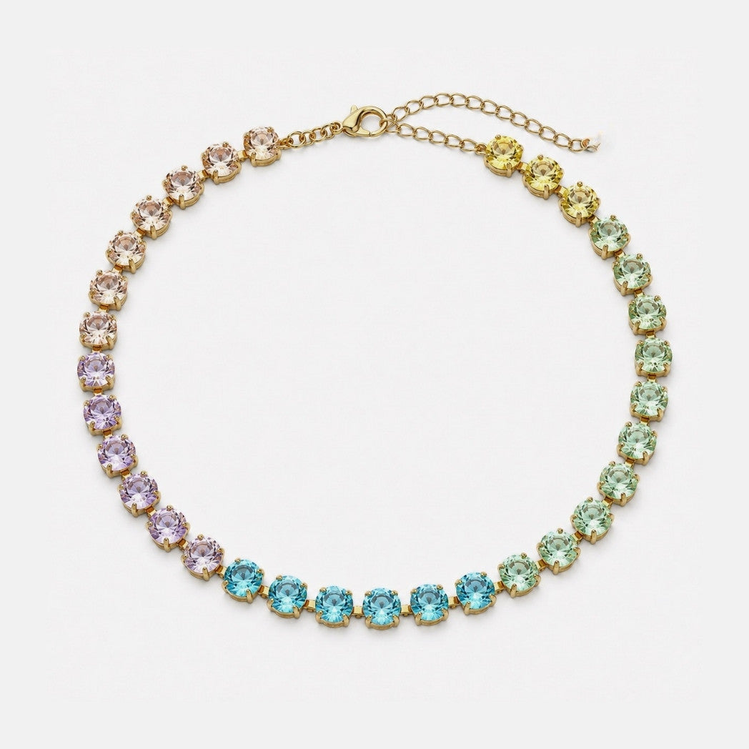 A gold chain necklace featuring a graduated row of round-cut clear crystals in pastel rainbow colors (pink, purple, blue, green, yellow) on a light gray background.