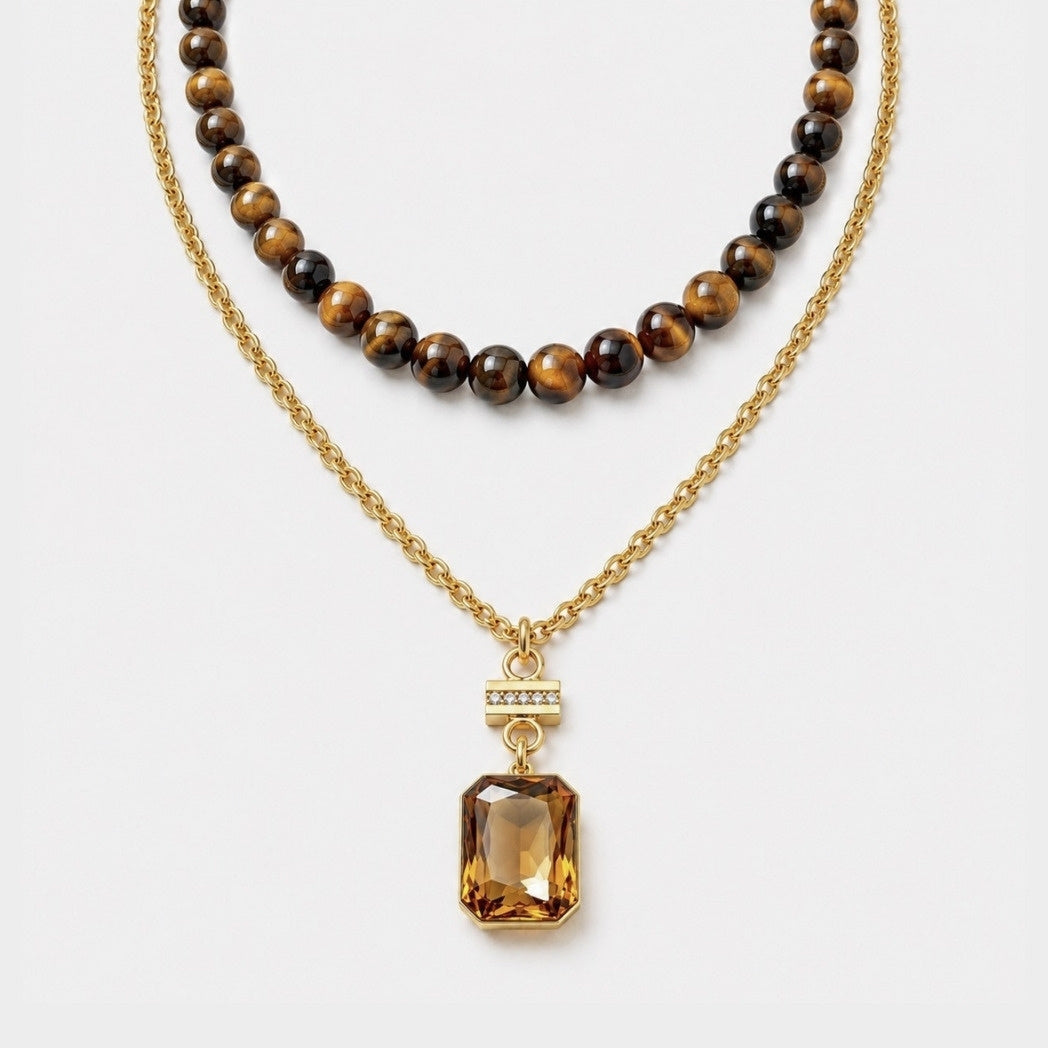 A double-strand necklace featuring a gold chain with a faceted amber gemstone pendant and a second strand of polished tiger's eye beads, displayed on a clean white background.