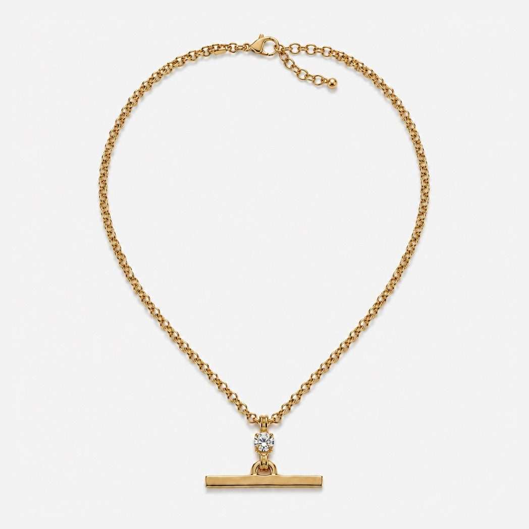 A gold chain necklace with a horizontal bar pendant and a small clear gemstone accent, displayed on a light gray background.