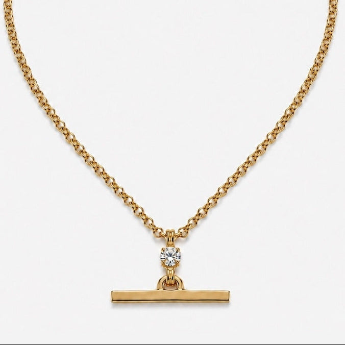 A gold chain necklace with a horizontal bar pendant and a small clear gemstone accent, displayed on a light gray background.