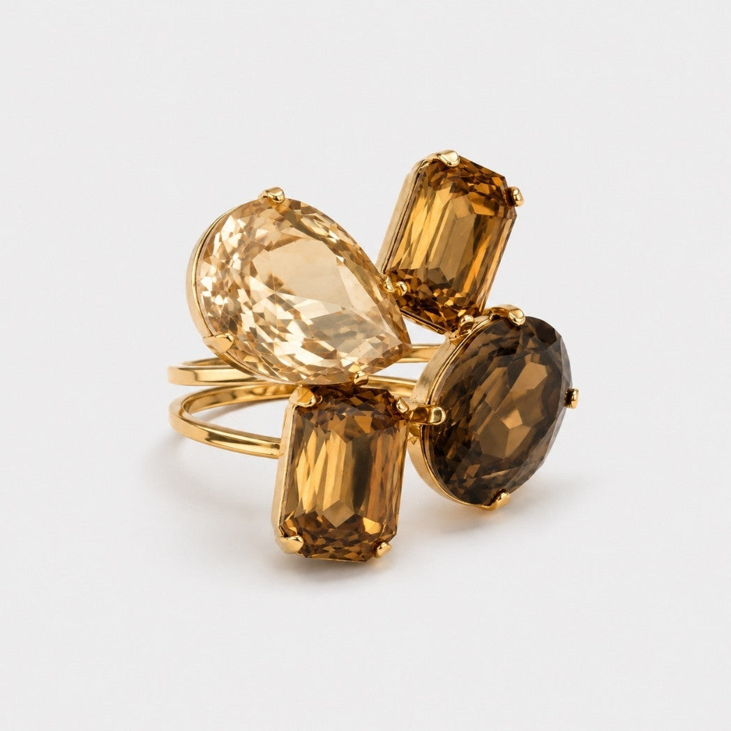 A gold ring featuring four large, faceted gemstones in different shapes and tones: a pear-cut light amber, a rectangular amber, an oval smoky brown, and a rectangular champagne gold.