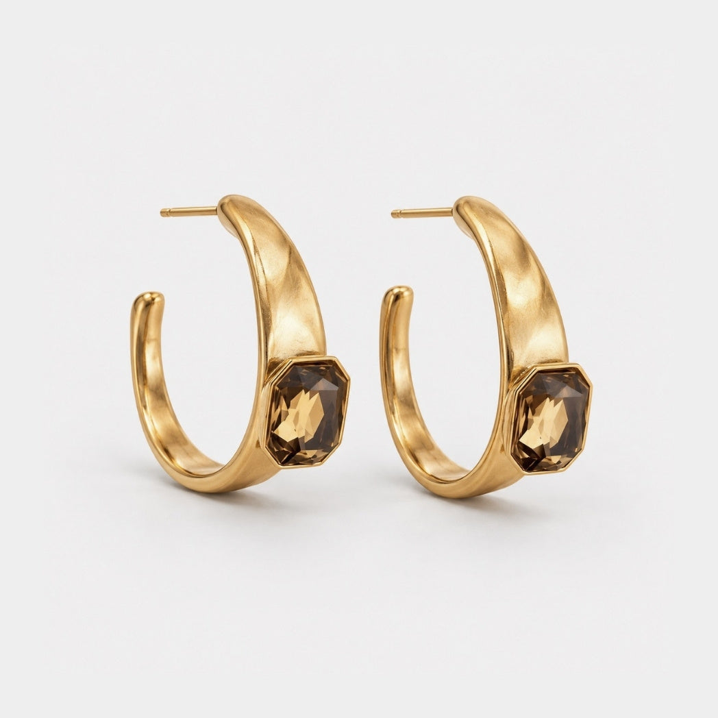 A pair of gold hoop earrings with a textured finish and a faceted dark brown gemstone set on the front, shown against a light gray background.