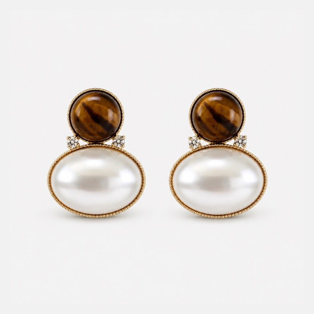 A pair of gold drop earrings featuring a round brown tiger's eye stone on top of a larger oval white pearl, accented with small clear stones, set against a light gray background.