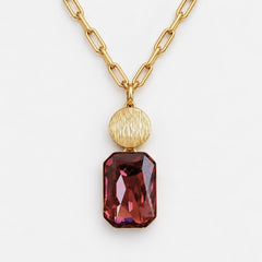 A gold paperclip chain necklace featuring a textured gold oval pendant and a faceted rectangular dark red gemstone pendant, shown on a light gray background.