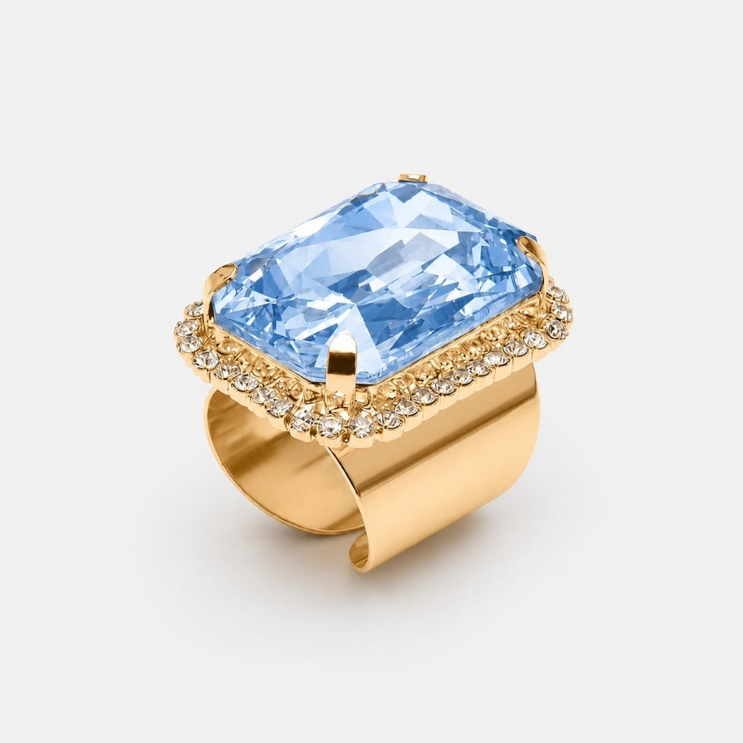 A gold ring featuring a large, cushion-cut light blue gemstone surrounded by a halo of small clear stones, set on a wide polished gold band, shown on a light gray background.