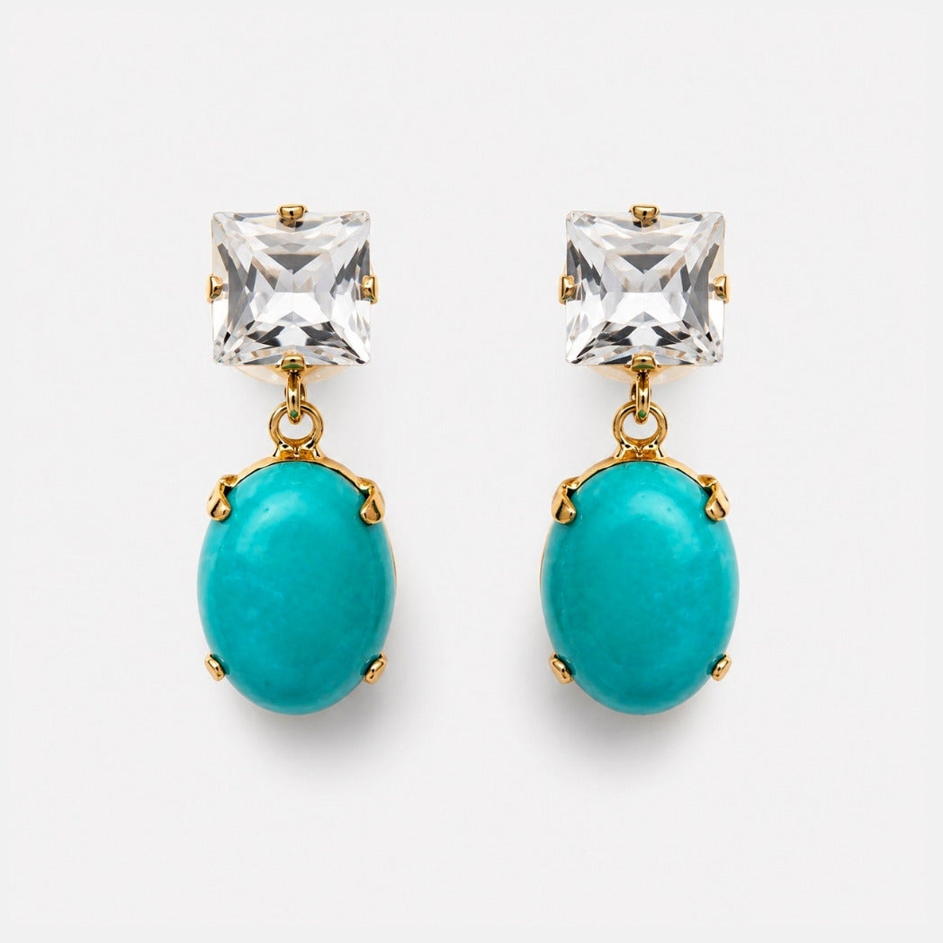 A pair of gold-tone drop earrings featuring a clear square-cut crystal and an oval turquoise stone, set against a plain light gray background.