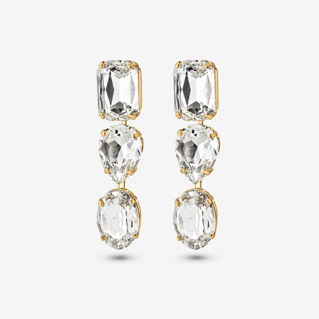 A pair of gold drop earrings featuring three clear faceted gemstones in rectangular, pear-shaped, and oval cuts, set against a light gray background.