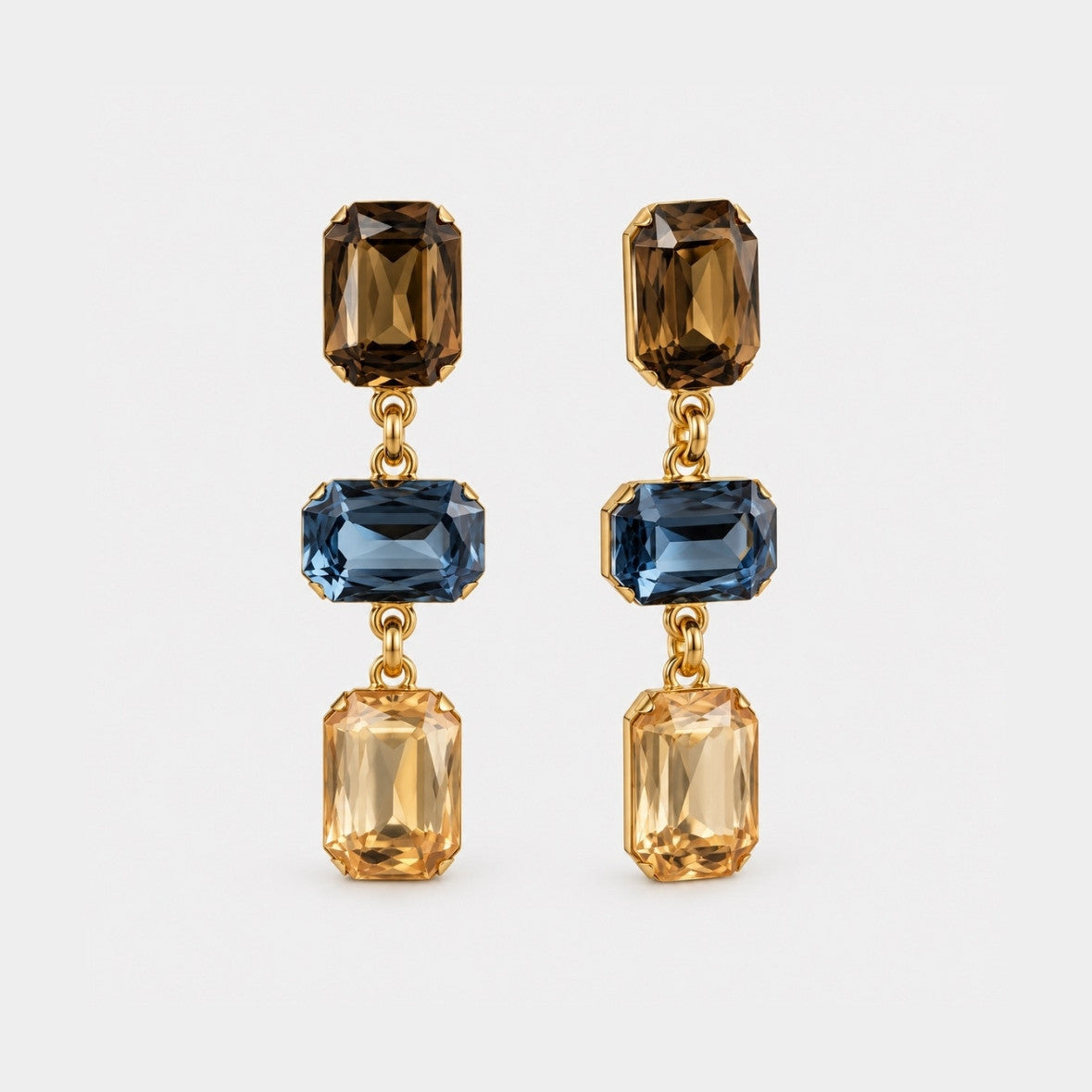 A pair of gold drop earrings featuring three rectangular faceted gemstones in each earring: a dark brown top stone, a blue middle stone, and a light amber bottom stone, shown on a light gray background.