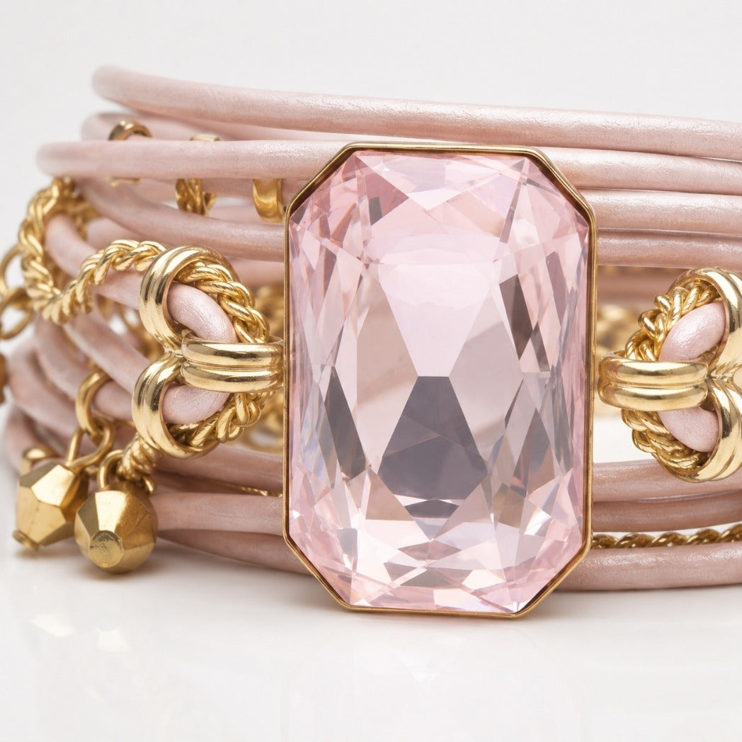 A close-up of a multi-strand bracelet featuring pale pink faux leather bands, gold rope details, and a large faceted pink gemstone centerpiece in a gold-toned bezel.