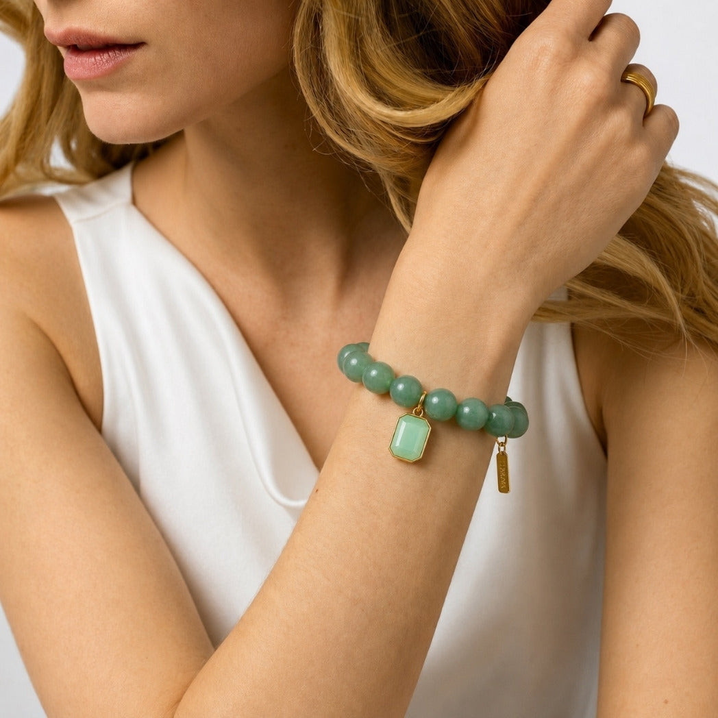A close-up of a woman wearing a green beaded bracelet with a rectangular mint-green stone charm and a small gold tassel, paired with a white sleeveless top.
