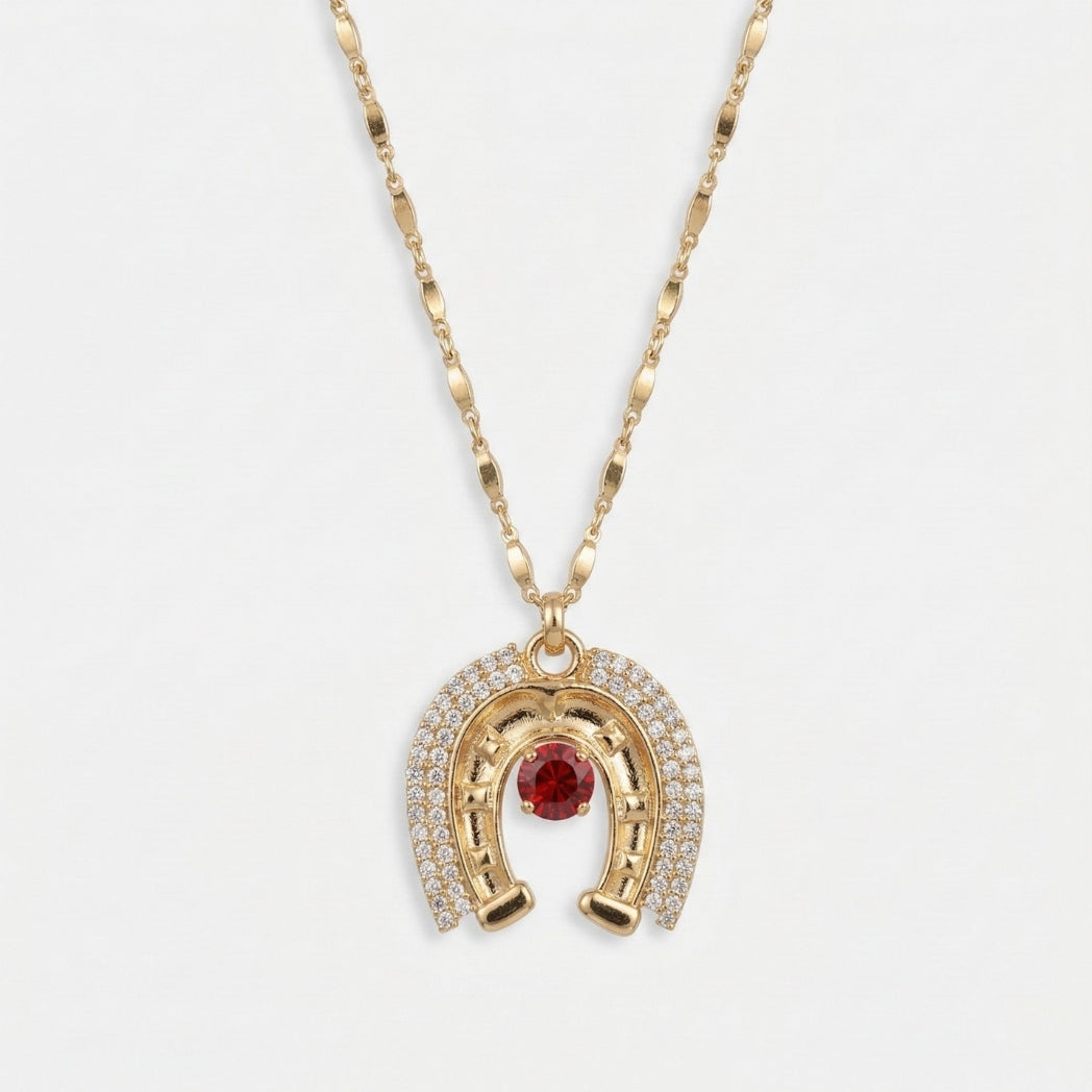 Gold necklace with a horseshoe pendant featuring a red gemstone on a white background