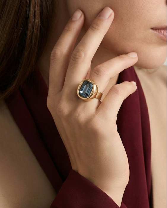 Close-up of a hand wearing a gold ring with a blue gemstone, against a neutral background.