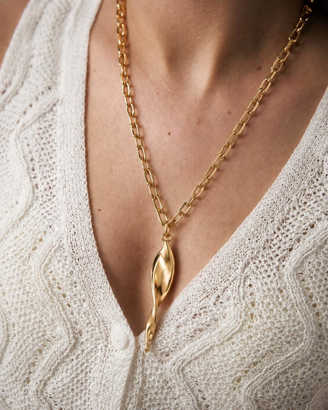 Gold chain necklace with a leaf pendant on a person wearing a white textured top.