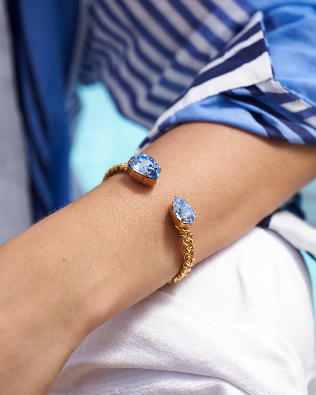 A close-up of a person's arm wearing two gold cuff bracelets, each featuring a light blue teardrop-shaped gemstone. The person is wearing a blue and white striped top and a white shirt.