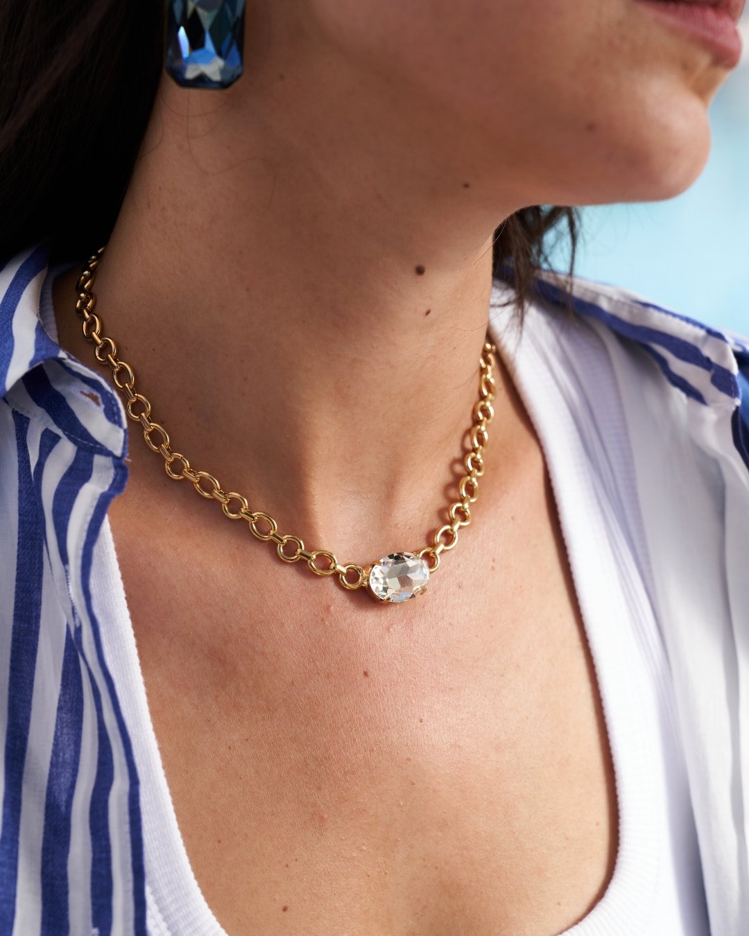 A close-up lifestyle shot of a woman wearing a gold chain necklace with a clear faceted oval gemstone pendant, styled with a blue and white striped shirt.