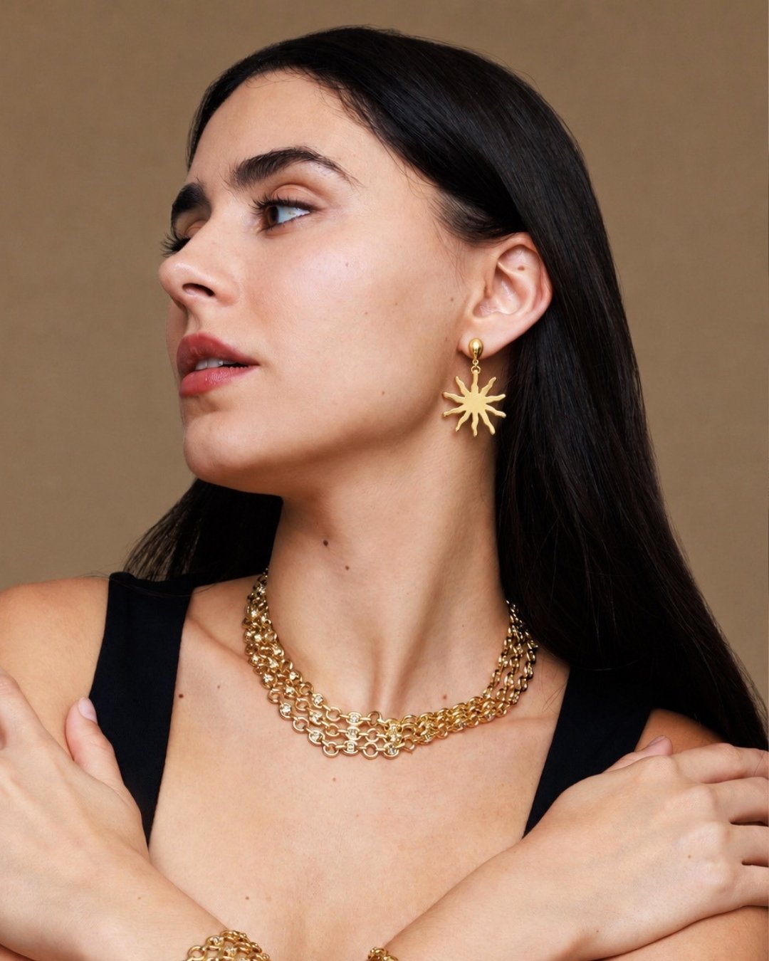 Woman wearing gold jewelry including earrings and a necklace against a brown background
